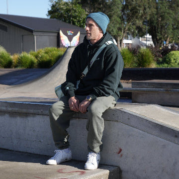 Be Better Owner Trent, sitting on a concrete skate ledge with trees and a building in the background .
Wearing the Heavy Mens Crew and Relax fit Track Pants and a Cuff Beanie with the Be Better Duffel Bag in Mint Green over his shoulder  a gold guess watch,with blue dial and some crisp white lacoste mens tennis shoes with the wee green croc 