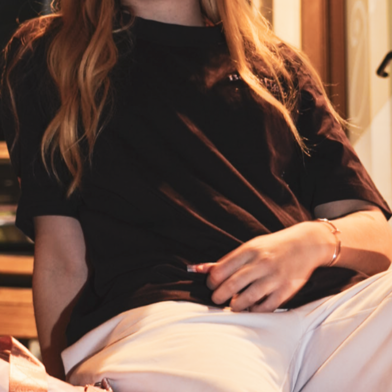 Woman sitting outside shops at night with a pink bag wearing be better pink track pants and a heavy Plum Purple tee with Dusty Pink Embroirered Be Better logos